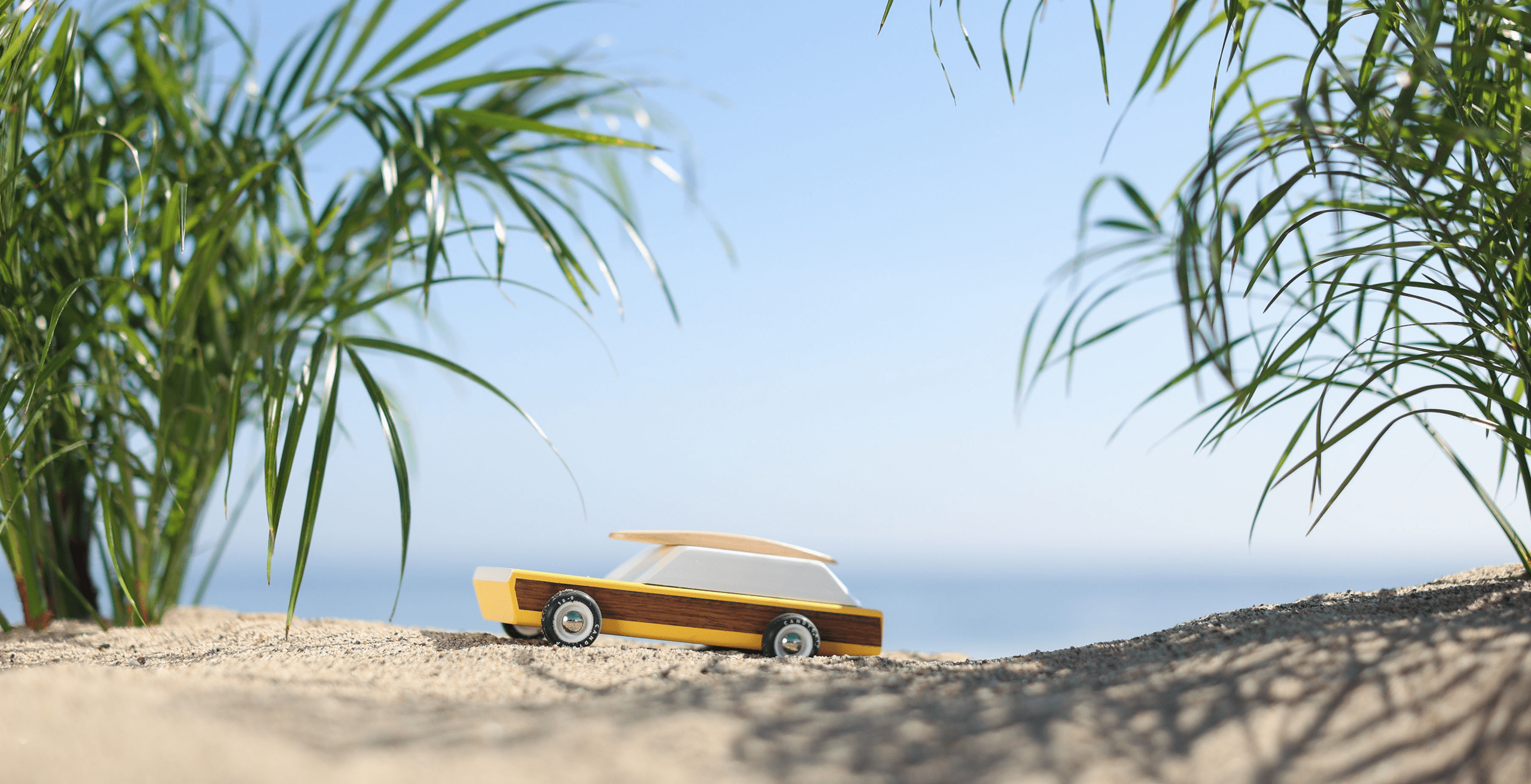 A Candylab wooden toy car sits on sandy ground, surrounded by green plants, with a clear blue sky in the background.