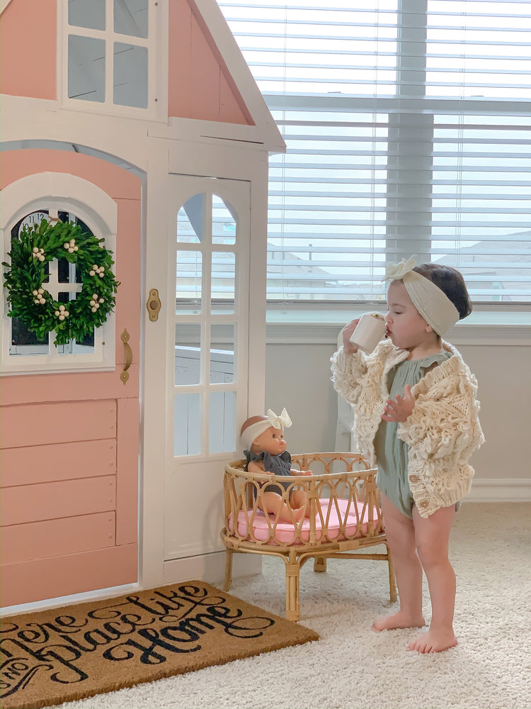 A toddler wearing a headband and knitted sweater drinks from a cup beside a pink and white playhouse in the cozy playroom. Soft toys are scattered around, while a poppie toys Poppie Crib Classic Collection sits on display nearby, with a doll comfortably resting inside. Light streams in from the window behind them.