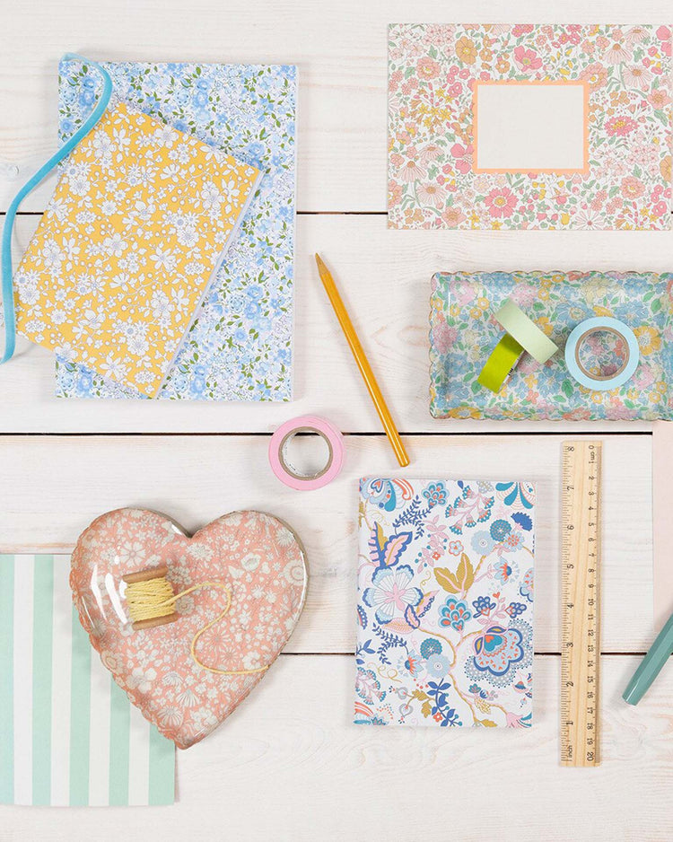 A flat lay of the meri meri x liberty heart small accessory tray by meri meri, styled with Liberty floral print stationery, a pencil, ruler, ribbon, washi tape, and string on a white wooden surface.