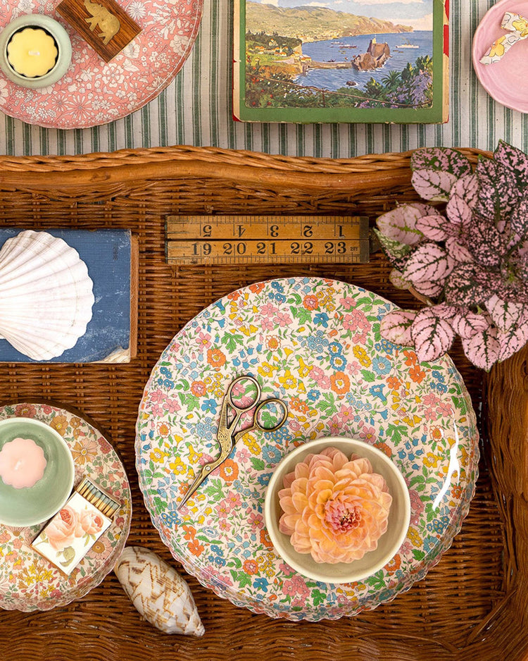 A meri meri x liberty round large accessory tray by meri meri holds scissors, a bowl with a peach dahlia, seashells, a candle, a ruler, a potted pink plant, and a vintage book atop a striped tablecloth.