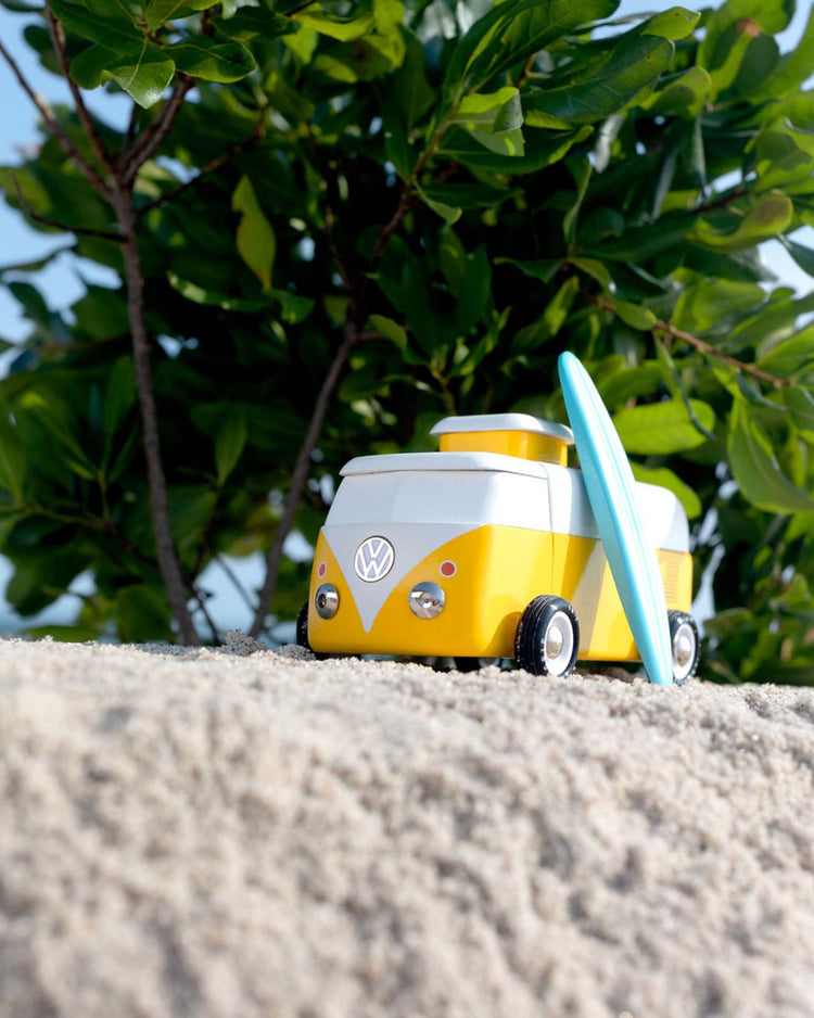 The candylab VW beach bus sunset, a small yellow and white toy van with a blue surfboard, sits on sandy ground with green leafy plants in the background.