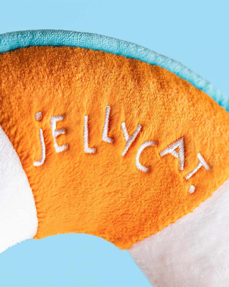 Close-up of the Jellycat amuseables life ring plush toy, featuring bright orange and white fabric with "JELLYCAT" embroidered in white on the orange section, set against a light blue background.