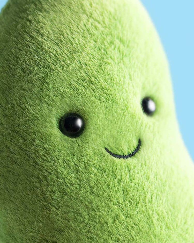 A close-up of the Jellycat amuseables soyella edamame bean plush toy with a soft, fuzzy texture, black button eyes, and a small smiling mouth against a light blue background.