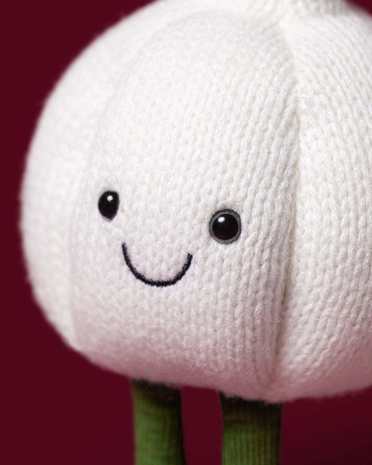 A close-up of the Jellycat Amuseables Garlic plush toy, featuring a knitted white garlic bulb with a cheerful smile, black eyes, and soft green stalk legs, set against a dark red background—an ideal whimsical accent for your next Halloween party.