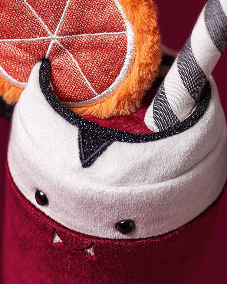 A cuddly Amuseables Vampop Drink by Jellycat, featuring vampire fangs and eyes, a soft orange slice topper, and a black-and-white striped straw—all set against a burgundy background—makes a perfect playful plush or gift for Halloween.