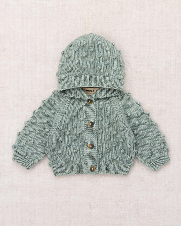 A light green ("onsen") baby hooded popcorn cardigan by misha + puff, hand-knit from soft Merino wool with a front button closure and a textured bobble stitch pattern, is laid flat on a pale, textured background.