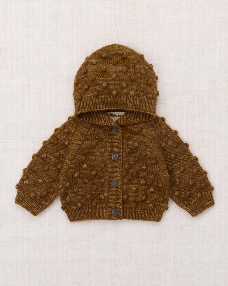 The baby hooded popcorn cardigan in nutmeg by misha + puff, crafted from hand-knit Merino wool with front buttons and a charming raised dot stitch pattern, is displayed laid flat on a light fabric background.