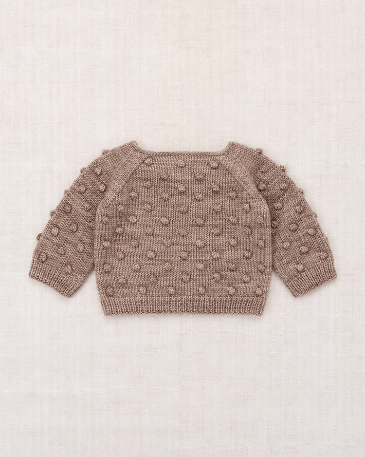 The misha + puff baby popcorn sweater in turtledove is a hand-knit, long-sleeved baby sweater made from soft Merino wool, featuring a charming bobble popcorn stitch pattern on the front, back, and sleeves—beautifully displayed flat on a beige background.