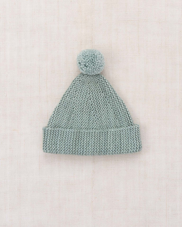 A light gray "baby garter hat in onsen" by misha + puff, featuring a knitted Merino wool design with a folded, adjustable brim and a large pom-pom on top, displayed on a neutral textured fabric background.