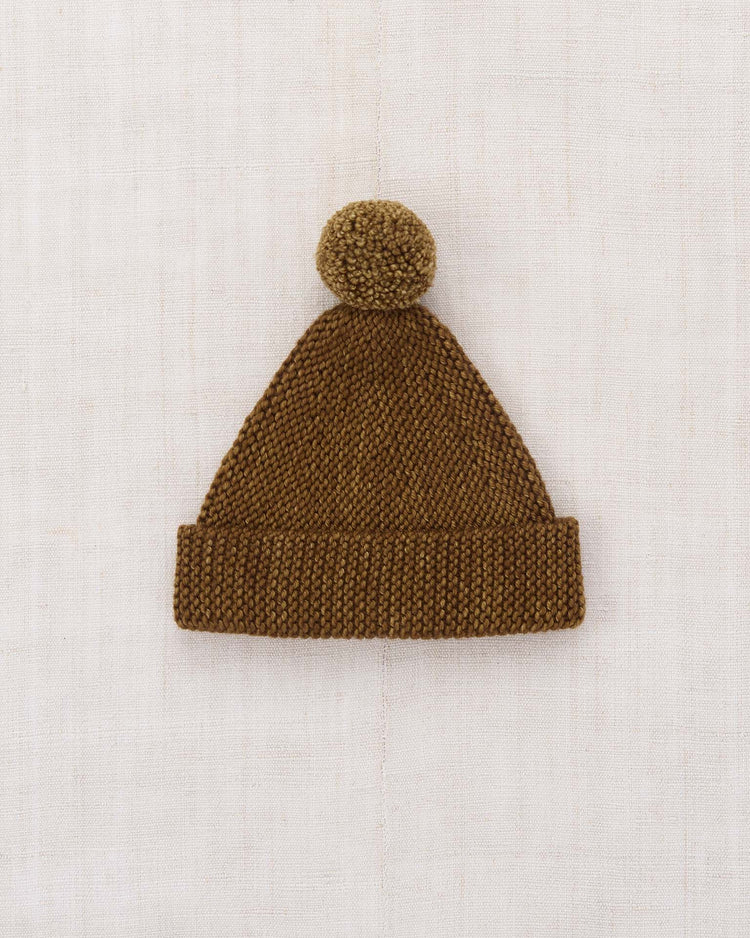 The baby garter hat in nutmeg by misha + puff, featuring a folded brim and a large pom-pom on top, is laid flat on a light, textured fabric background.