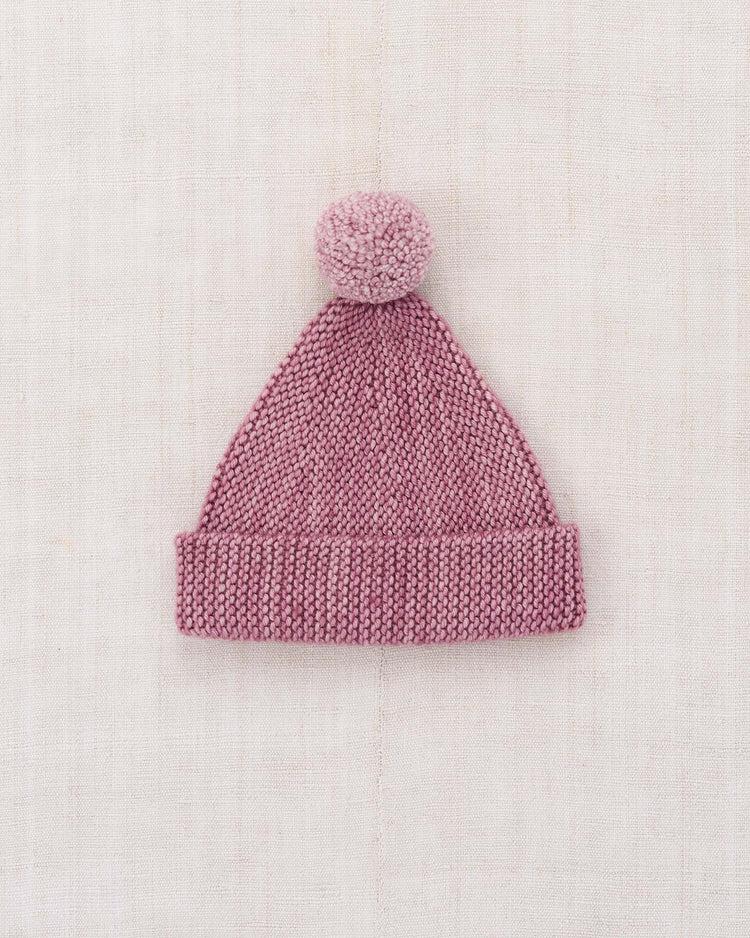 The "baby garter hat in antique rose" by misha + puff, featuring a knitted design with a folded adjustable brim and a large pom-pom on top, is displayed on a light, textured fabric background.
