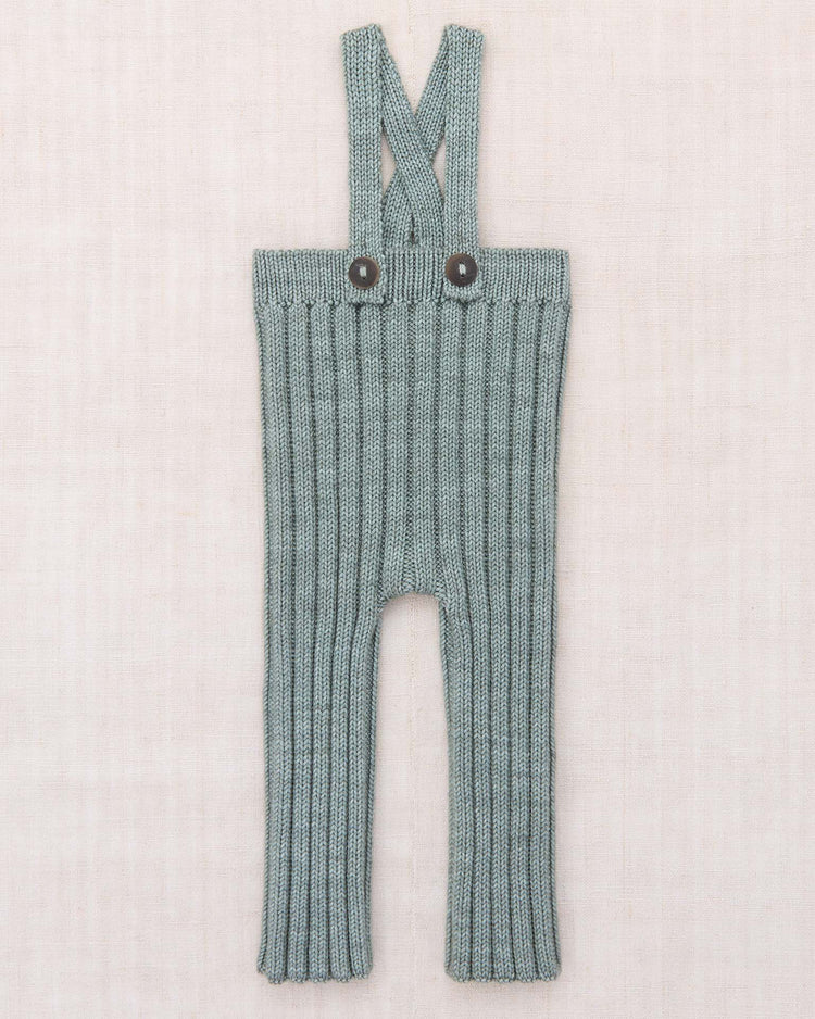 The misha + puff baby rib longies in onsen, crafted from soft merino wool knit with a gray ribbed texture, feature crossed adjustable shoulder straps and two dark buttons, shown laid flat on a light beige fabric background.