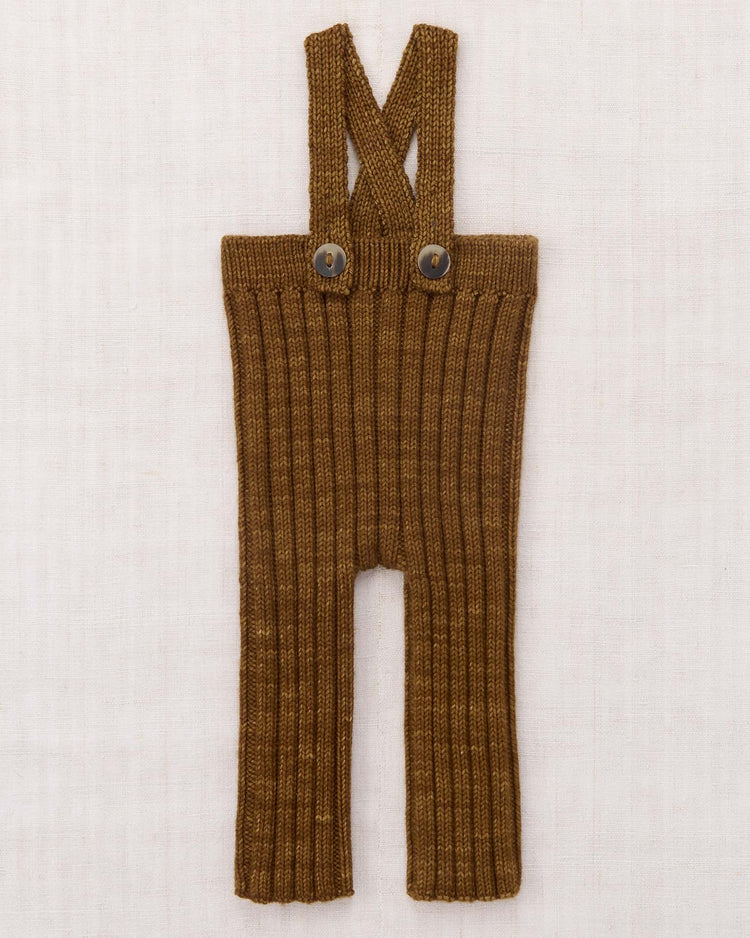 The misha + puff baby rib longies in nutmeg, featuring a brown ribbed knit and adjustable crisscross straps with two front buttons, are laid flat on a light, textured background.