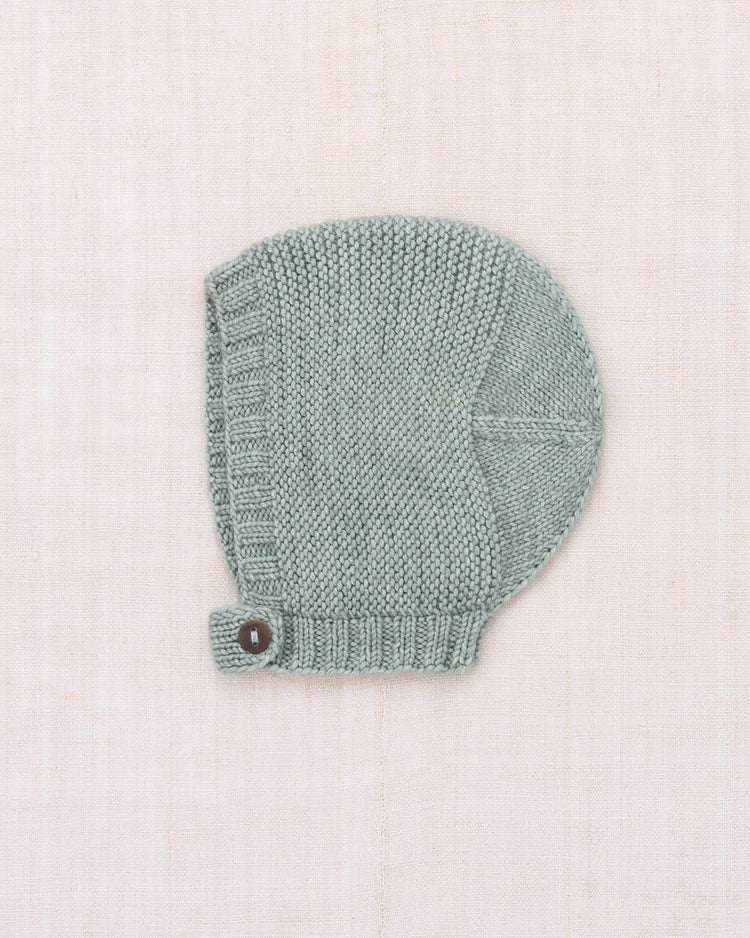 The baby beachwalk bonnet in onsen by misha + puff—a hand-knit light gray Merino wool bonnet featuring a horn button strap closure—is shown displayed flat on a pale beige textured background.