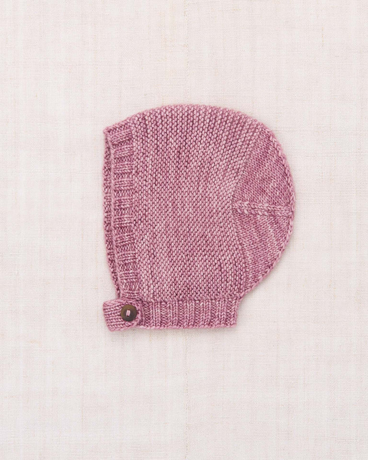 The baby beachwalk bonnet in antique rose by misha + puff is hand-knit from soft Merino wool and features a charming horn button closure on its strap, displayed lying flat on a light beige fabric background.