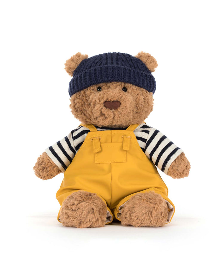 Jellycat's Bartholomew Bear Seafarer Outfit features the plush bear dressed in a navy blue beanie, black and white striped shirt, and bright yellow overalls, perfect for nautical adventures against a plain white background.