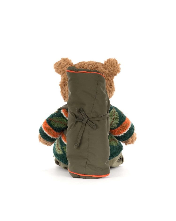 The Jellycat Bartholomew Bear Hiking Outfit features Bartholomew Bear dressed in a green, orange, and cream striped sweater with a large green sleeping bag tied on his back, all set against a plain white background.