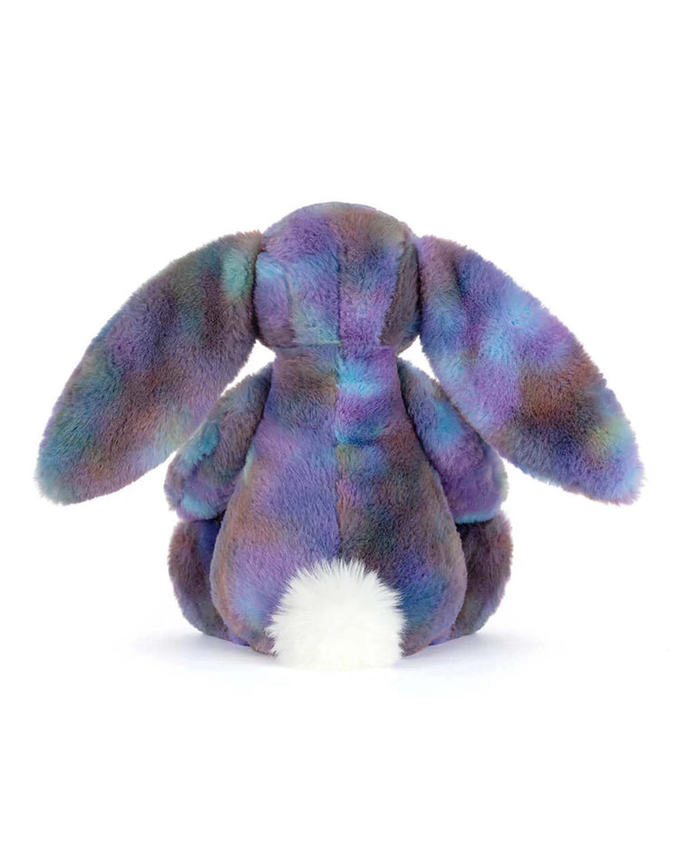 The Jellycat Zodihop Luxe Bunny is shown from behind, highlighting its long floppy ears, fluffy white tail, and plush fur in purple, blue, and green cosmic patterns against a plain white background.