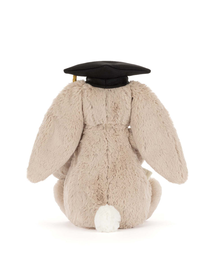 The Jellycat bashful bunny graduation outfit features a plush bunny facing away, dressed in a black cap with a gold tassel. Its floppy ears and white tail make it a charming graduation gift, shown on a plain white background.