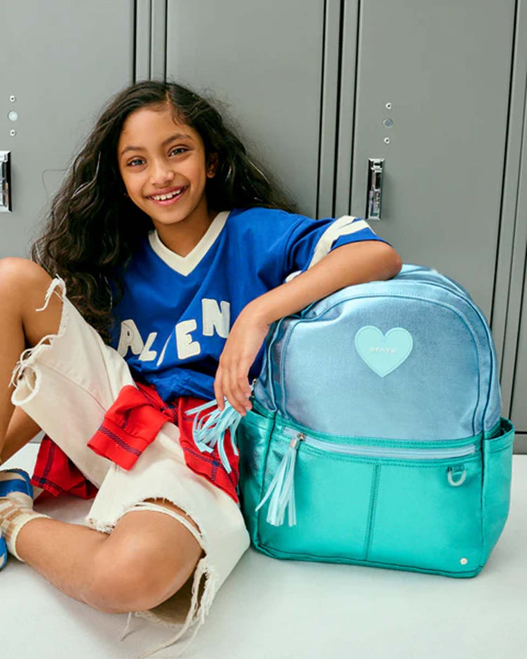 A smiling girl in a blue shirt and ripped jeans sits by gray lockers with a shiny blue + turquoise colorblock Kane Backpack from STATE Bags featuring a heart-shaped patch beside her.