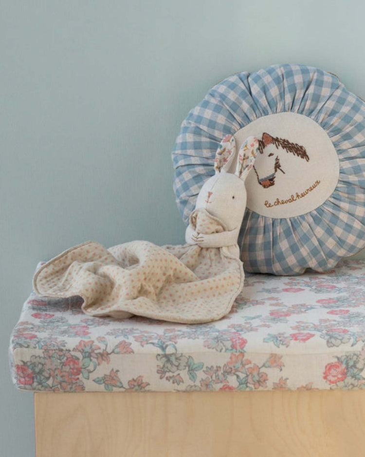 The lullaby friends, bunny, mini - nature by maileg sits with a cuddle cloth on a floral surface next to a blue gingham cushion with an embroidered horse and French text—an adorable baby gift against a light blue wall.