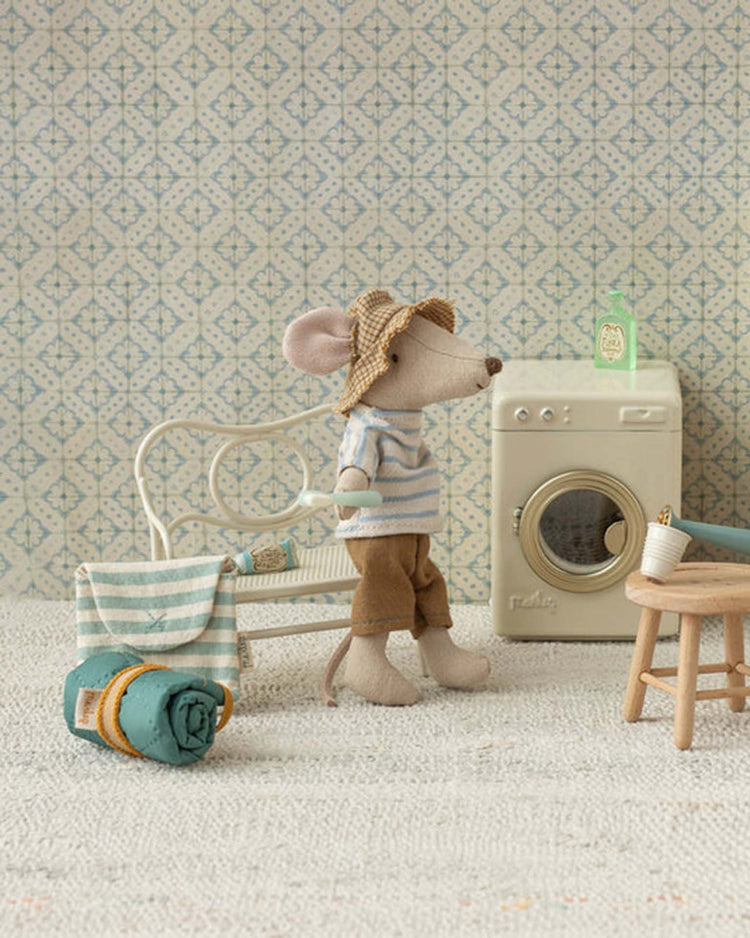 The maileg toothbrush set in bag, mouse – blue stripe features a small mouse toy dressed in a hat, striped shirt, and brown pants alongside a mini washing machine, white bench, and accessories, all in a cozy patterned room.