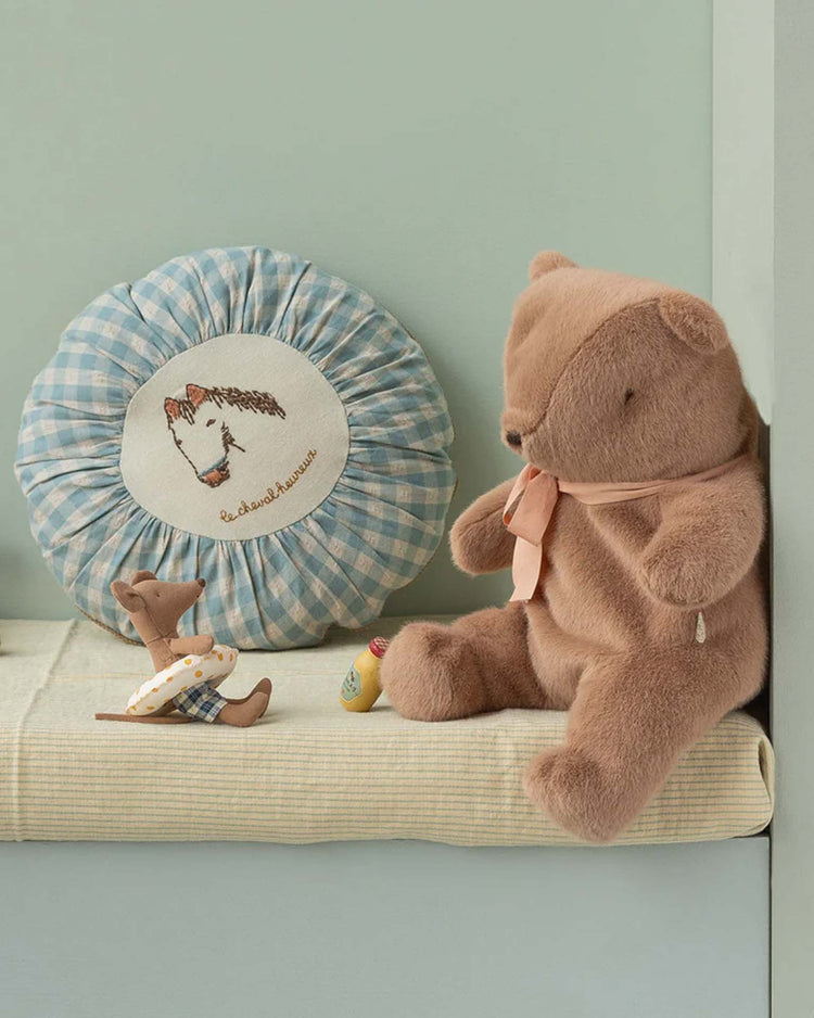 A soft brown medium teddy bear plush by maileg with a pink ribbon sits on a striped cushion next to a blue gingham pillow with animal embroidery. In front, two dressed toy mice hold tiny props.