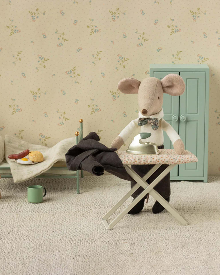 A maileg dad mouse wearing a wedding suit with a white shirt and bow tie stands at a tiny ironing board, pressing black pants in a cozy mini room featuring a bed, blanket, mug, food, and pale blue wardrobe.