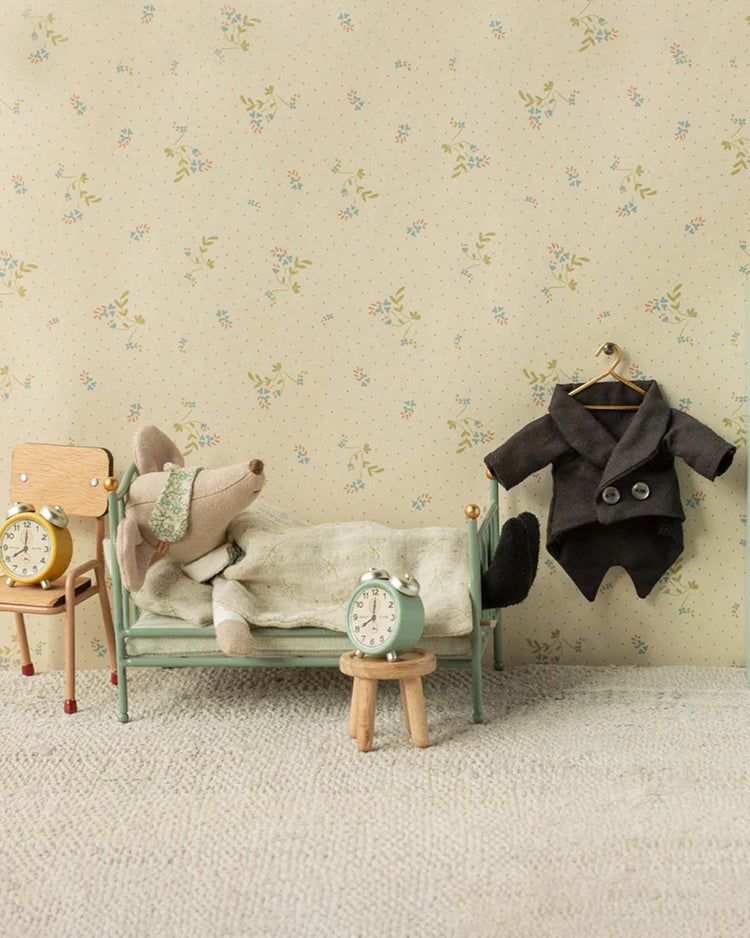 A Maileg dad mouse in a wedding suit lies under a blanket in bed. In the miniature room are two alarm clocks, a small stool, wooden chair, and a tiny coat hanging on the wall for extra charm.