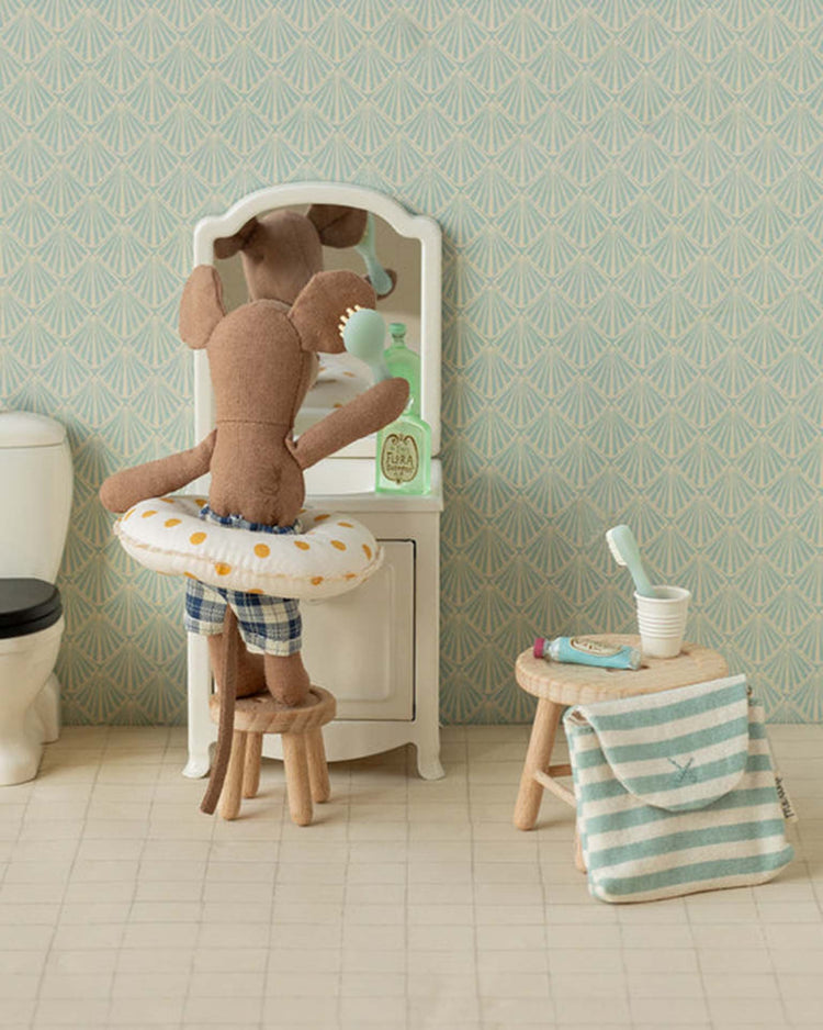 The Maileg toothbrush set in bag, mouse - blue stripe features a mouse in swim trunks with a float, brushing its teeth—ideal as a playset accessory or addition to a mouse family toy. Set includes patterned wallpaper, striped towel, and tiny toothbrush.