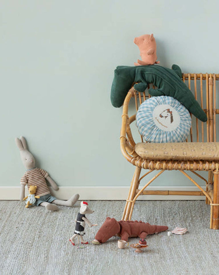 A rattan chair holds the Maileg small crocodile in vintage rose from Safari Friends and several plush toys, while a gray rabbit doll, yellow teddy, mouse doll, and toy tea set are on the floor. Some items are crafted with recycled polyester.