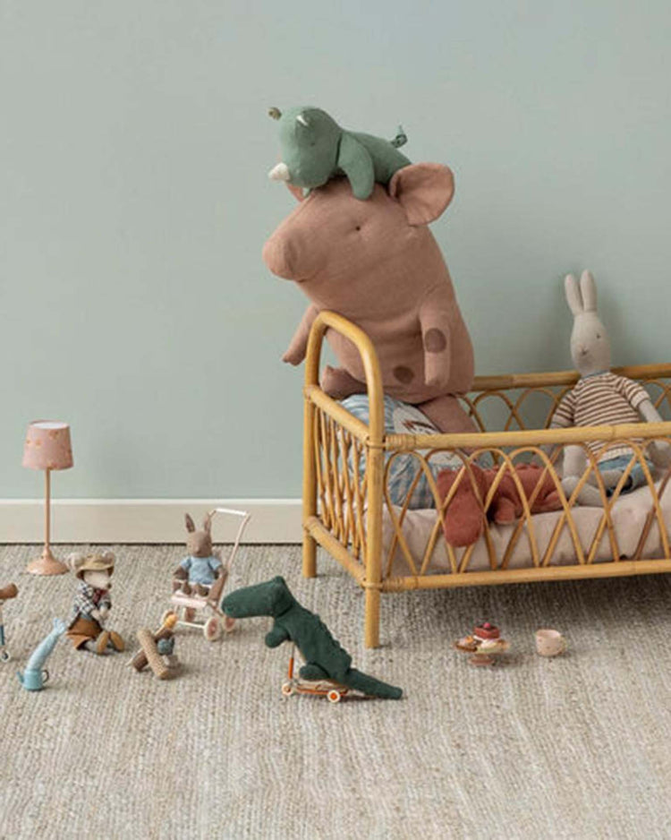 A cozy nursery features a rattan crib with plush toys, including the Maileg Safari Friends mini crocodile in forest green. More soft animals and wooden toys are scattered on the beige carpeted floor.