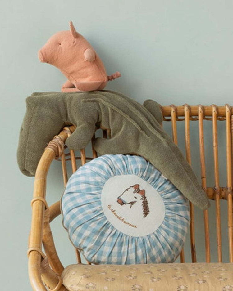 A wicker chair holds a round blue-and-white checkered pillow with a horse illustration, the maileg medium crocodile in olive green from safari friends, and a small pink plush pig on top.