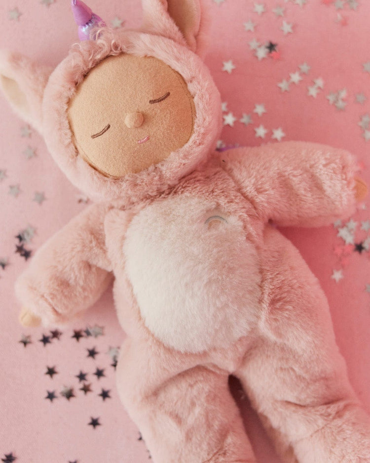 The cozy dinkum unicorn sugarpie pink by dinkum dolls is a soft, lovable plush toy in a pink unicorn outfit with closed eyes and a gentle smile, displayed on a pink surface accented with silver star confetti.