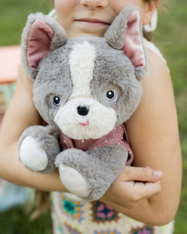 A child smiles while hugging a dinkum dolls dinkum dog gizmo plush toy with pink ears and blue eyes close to their chest outdoors.