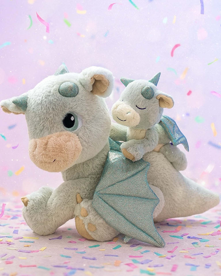 The dinkum dolls dinkum dragon blaze in green is a soft, light green plush dragon with sparkly wings, carrying a baby dragon on its back. Both have friendly faces and pastel details, set on confetti with a dreamy purple background.