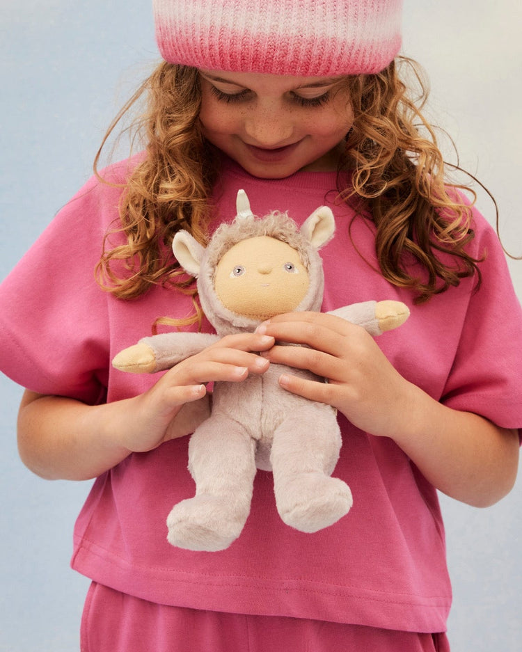 A child with curly hair, wearing a pink shirt and a pink-and-white beanie, smiles while holding the dinkum dolls Dinky Dinkum Doll Uma Unicorn Lilac, dressed in a soft light gray and lilac unicorn costume.