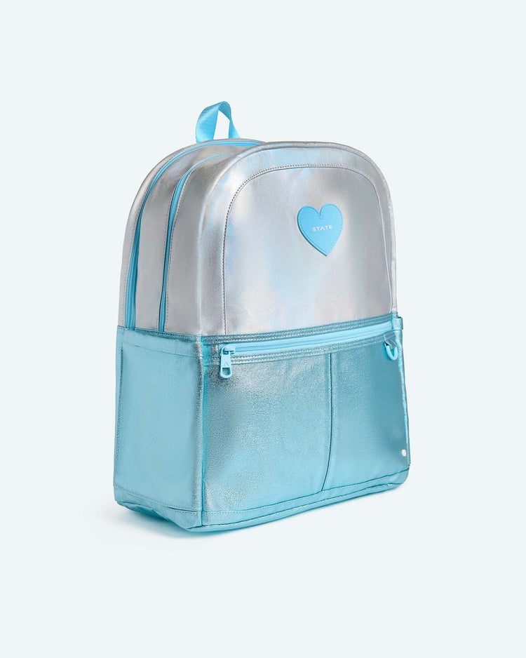 The Kane Large Double Pocket Backpack in Blue Iridescent by State Bags features an iridescent silver and blue design with a front zipper pocket, side pockets, and a small blue heart patch on the upper front. This stylish school backpack is equipped with light blue straps and a top handle for easy carrying.