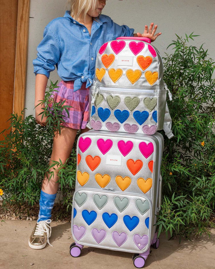 A person in a blue shirt and colorful shorts stands by suitcases and a STATE Bags Kane Backpack in Happy Fuzzy Hearts, surrounded by green plants.