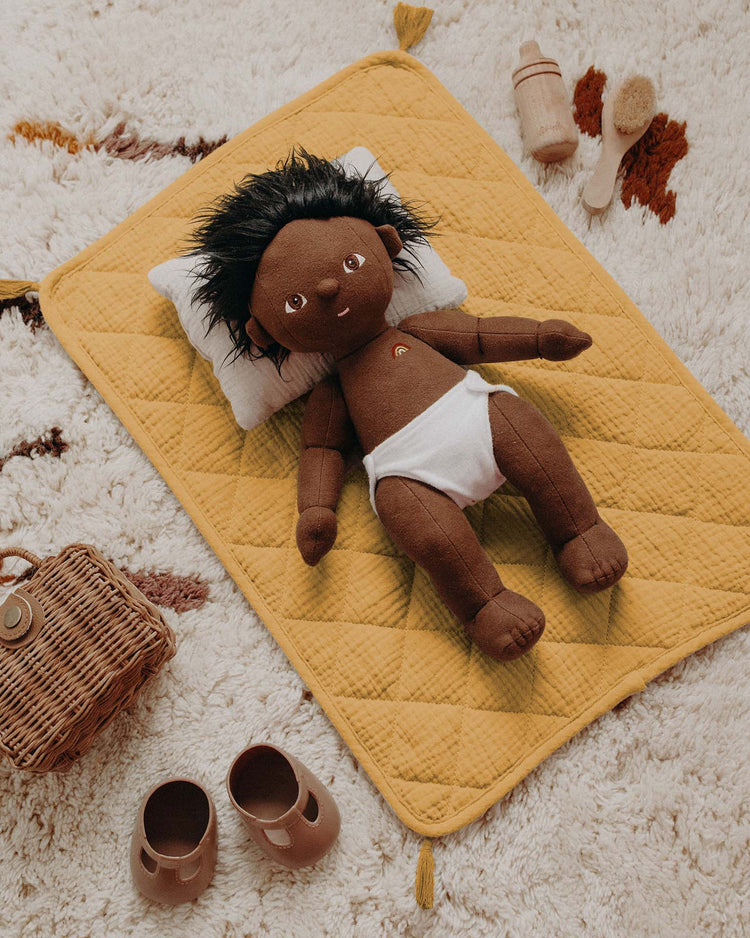 A dinkum dolls strolley bedding set in honey features a dark-skinned cloth doll with black hair, a white diaper, lying on a yellow quilted mat with pillow and cotton muslin blanket, surrounded by brown shoes and wooden toys on a cream rug.