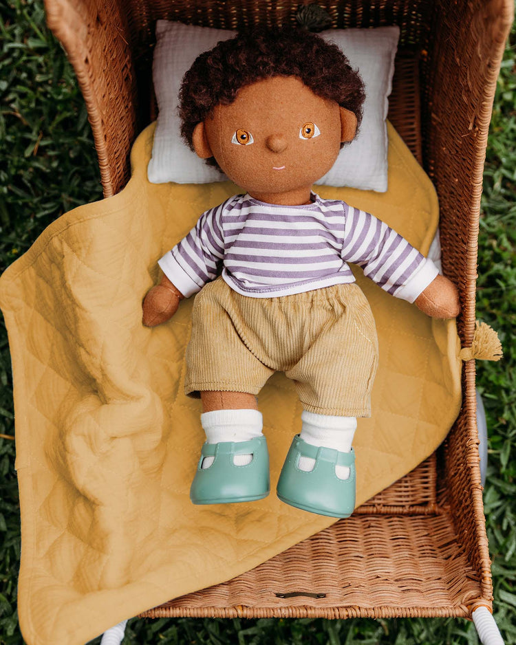 A soft cloth doll with brown skin, curly hair, and brown eyes rests in a wicker basket on green grass, covered by the dinkum dolls strolley bedding set in honey. The doll wears a striped shirt, tan shorts, white socks, and teal shoes.