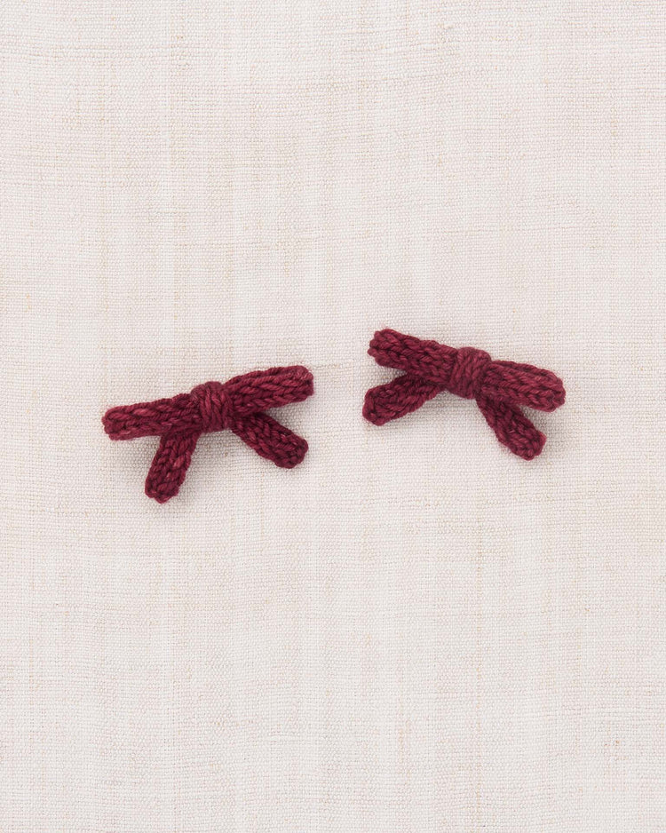Two small, maroon goldie bows from the misha + puff Cambridge set, hand-knit from soft Merino wool, are placed side by side on a light beige, textured fabric background.