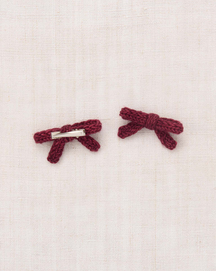Two small maroon hand-knit bows from the misha + puff goldie bow set in cambridge, crafted from soft merino wool, are placed on a light beige fabric background. One bow reveals the alligator clasp, while the other displays the charming front detail.