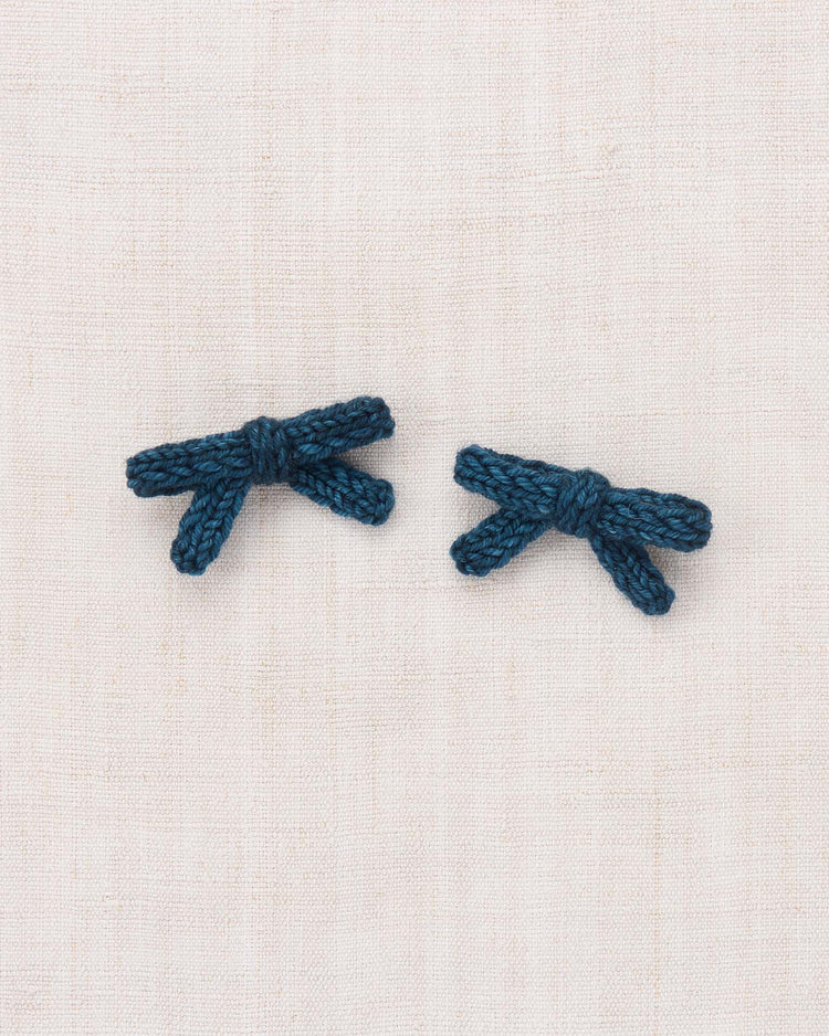 Two petite, marine blue bows from the misha + puff goldie bow set, hand-knit from soft merino wool, are displayed side by side on a light beige, textured fabric background.