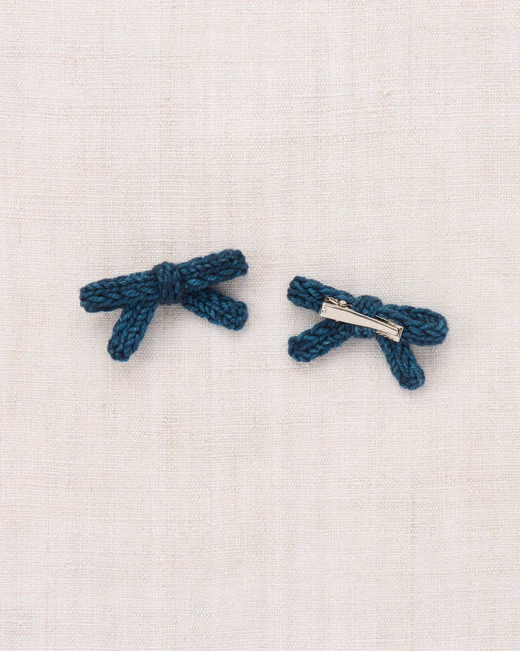 Two petite, marine blue Goldie Bow hair clips by misha + puff, hand-knit from soft merino wool, are displayed on a light beige fabric background. One clip highlights the graceful bow design, while the other reveals its secure alligator clip clasp on the reverse.