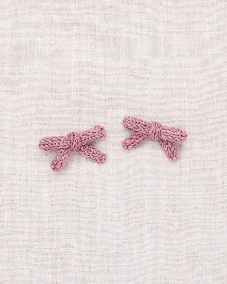 Two small, hand-knit bows from the misha + puff goldie bow set in antique rose are placed side by side on a light beige fabric background.