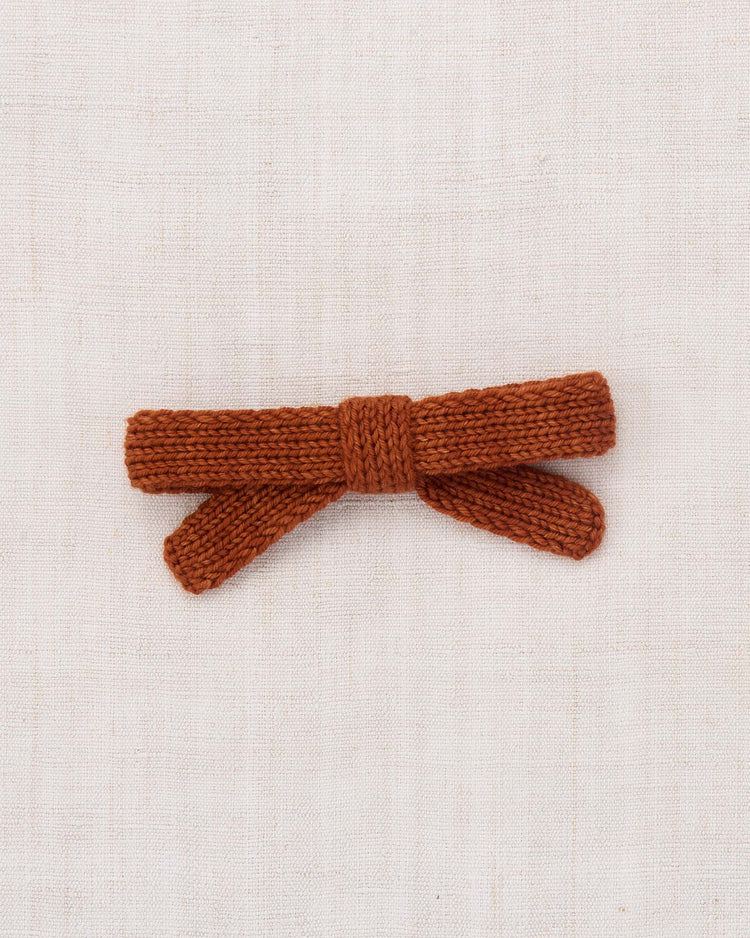 A hand-knit jumbo goldie bow in sabi by misha + puff lies flat on a light beige textured fabric background.