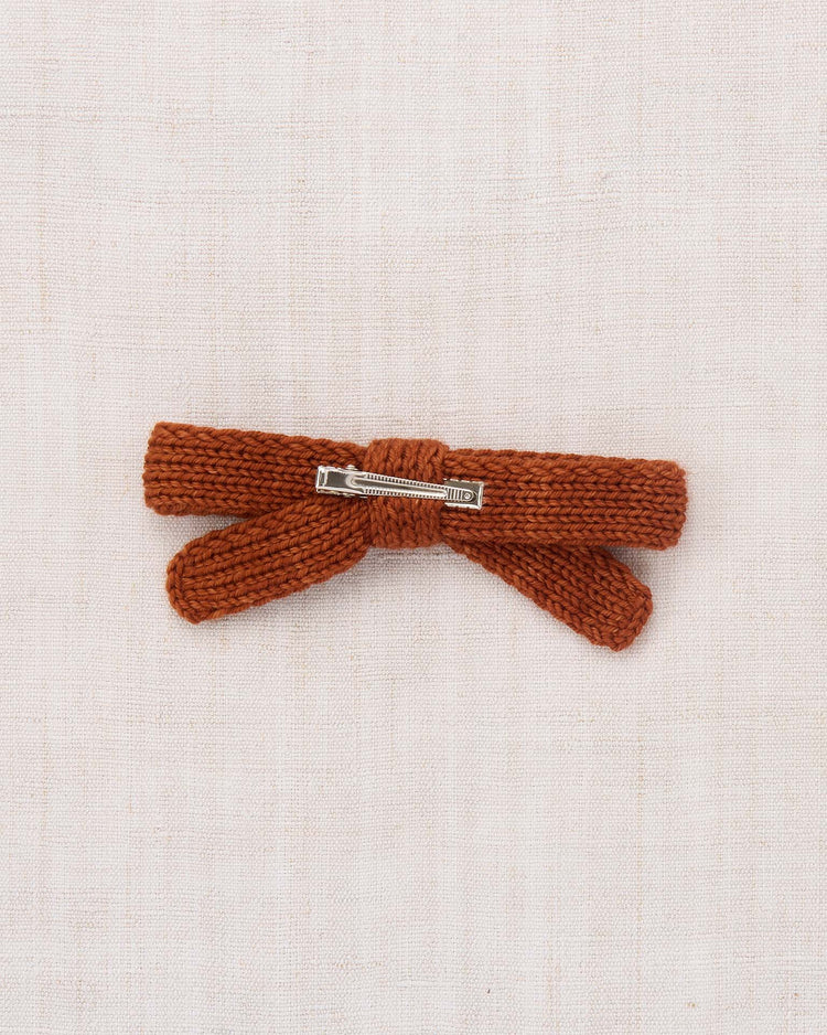 The jumbo goldie bow in sabi by misha + puff is a rust-colored, hand-knit hair clip made from soft merino wool and secured with a sturdy alligator clip, shown on a light beige fabric background.