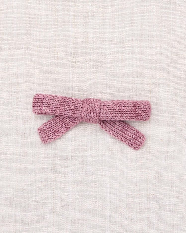 A jumbo Goldie bow in antique rose by misha + puff, hand-knit from soft merino wool, lies flat on a light, textured fabric background.