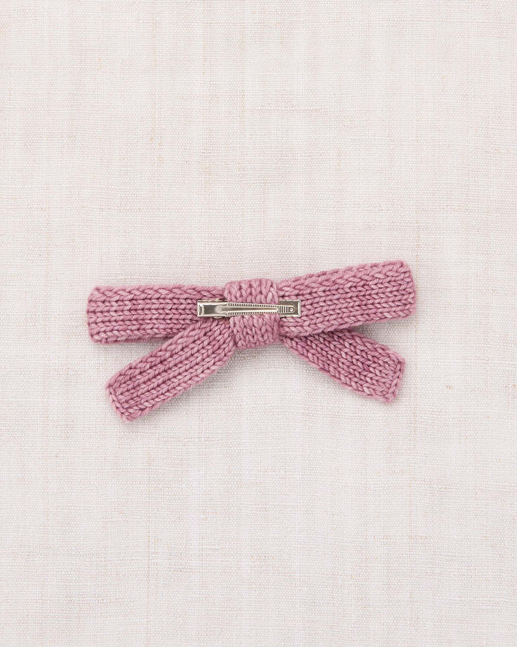 A jumbo goldie bow in antique rose by misha + puff, hand-knit from soft merino wool with a silver alligator clip, displayed on a light beige fabric background.